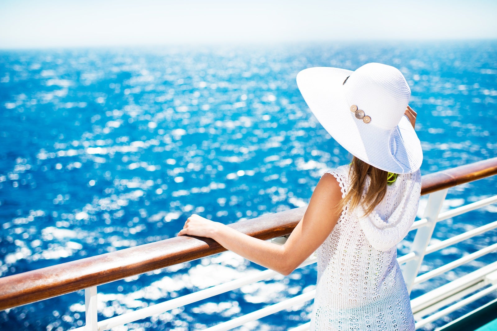 A woman in a white dress and white hat standing against a cruise ship railing with her back to the camera