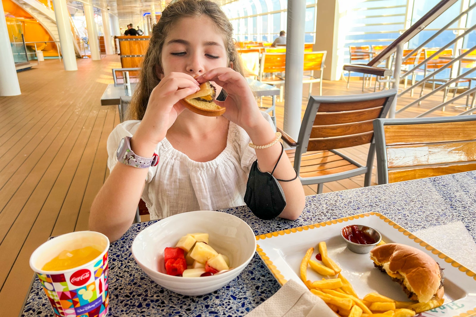 A young girl eating a burger and a bowl of fruit on a cruise ship 