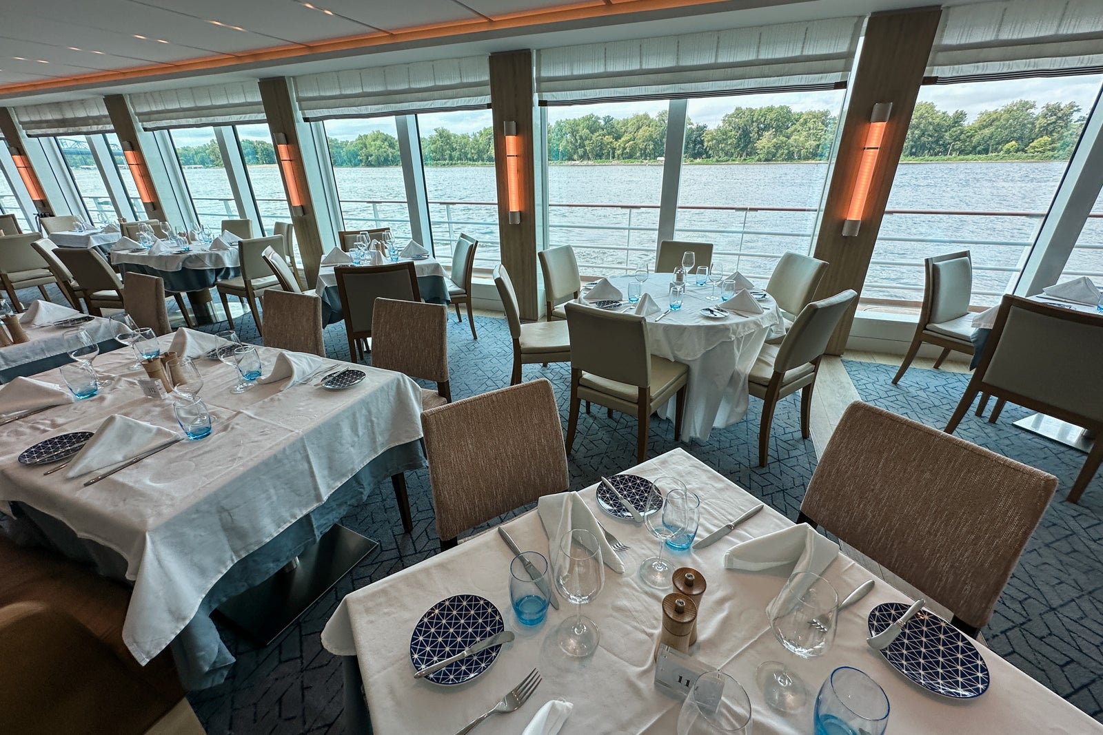 A cruise ship dining room where tables with white tablecloths are facing floor-to-ceiling windows