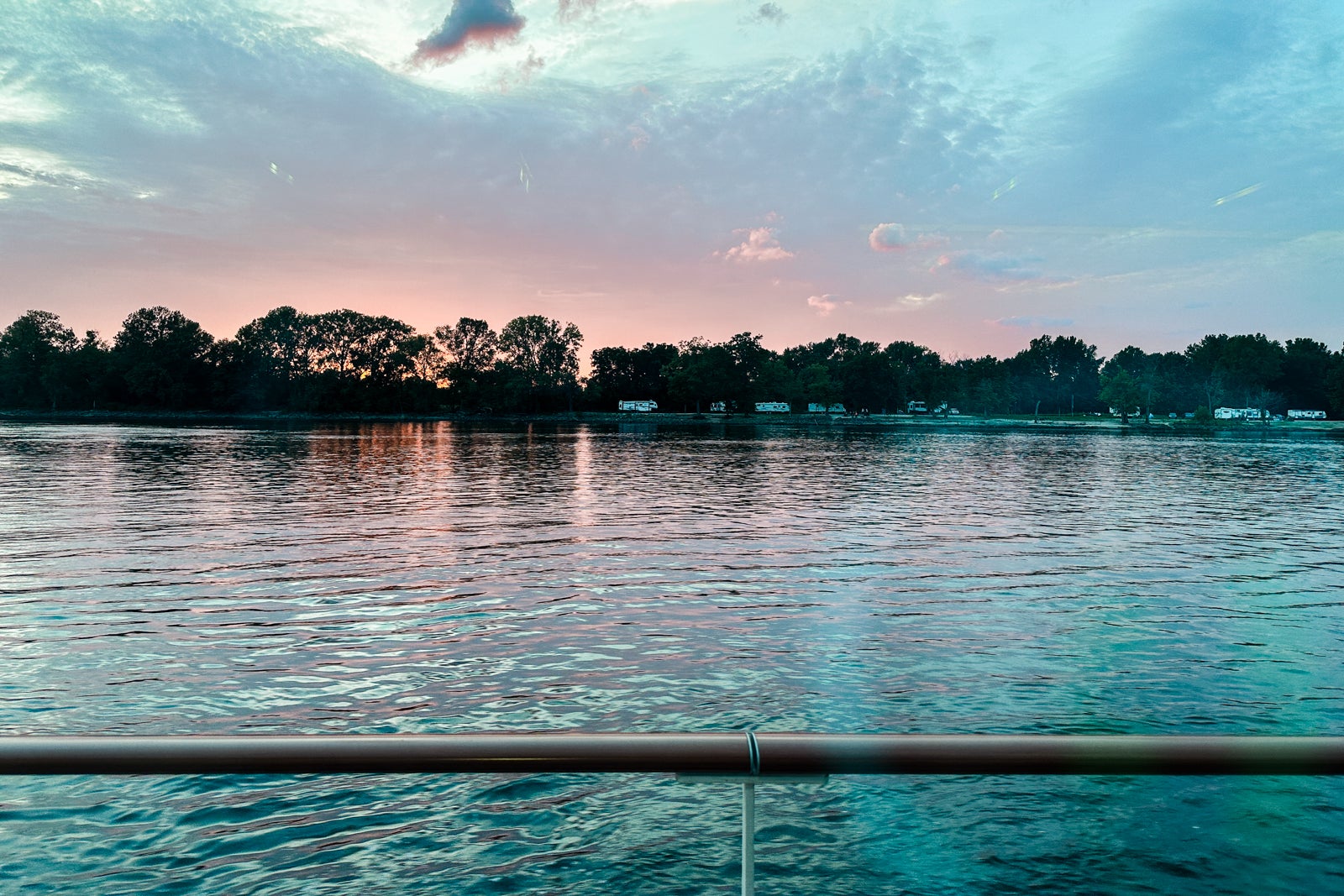 A cotton candy-colored sky at sunset above trees along the Mississippi River with a cruise ship railing in the foreground