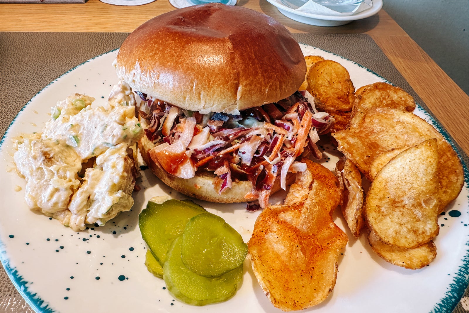 A white plate with a pulled-pork sandwich, potato salad, pickles and chips