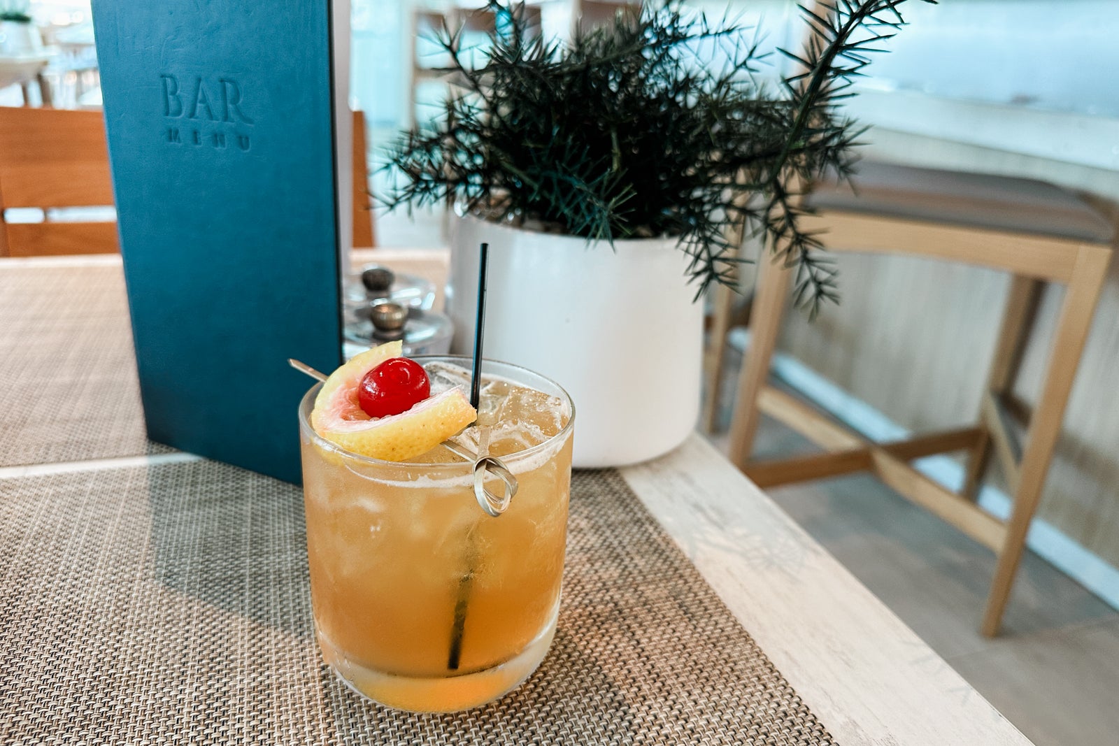 A yellow cocktail with a cherry garnish and a black straw sitting on a table in front of a plant and a drink menu