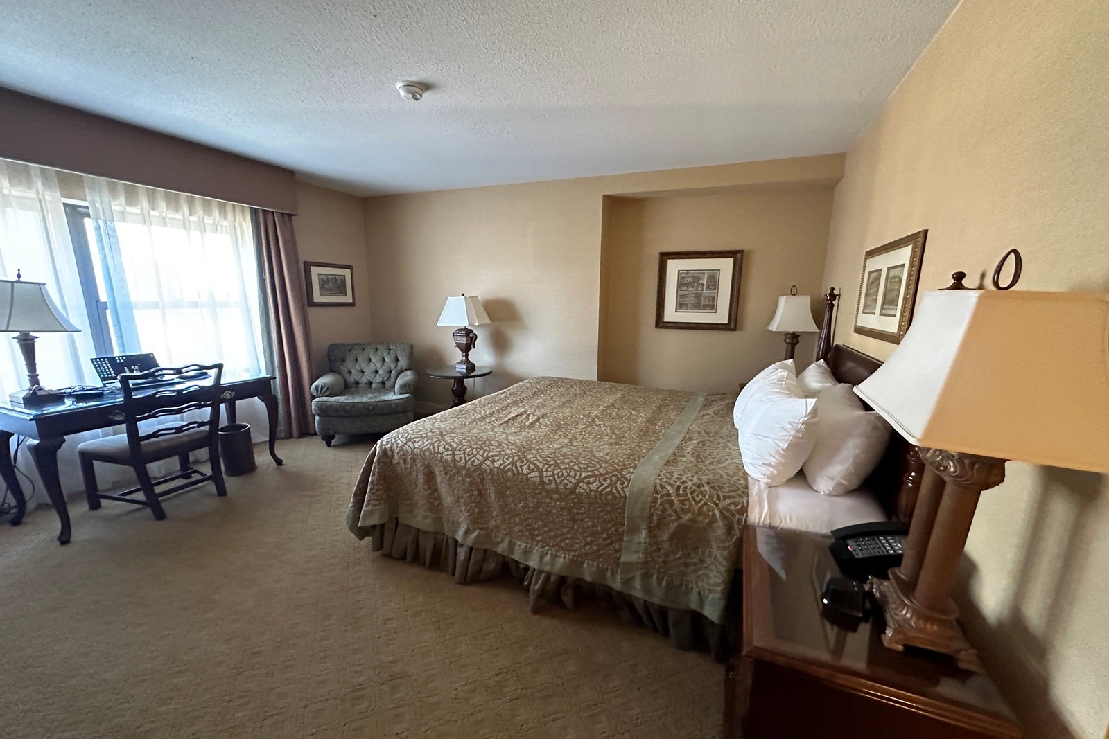 A hotel room with a queen bed, tan walls, nightstand, chair, lamps and desk