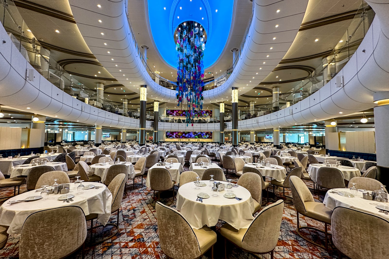 A view from the first floor of a cruise ship dining room with tables, chairs and a colorful glass sculpture overhead