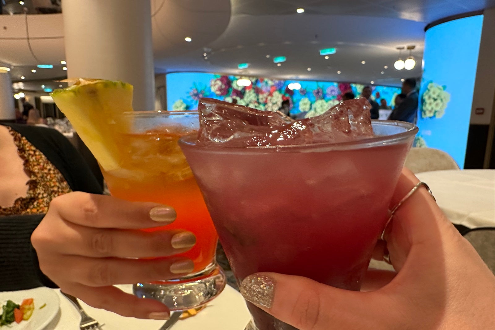 Two hands holding cocktails and toasting in a cruise ship dining room