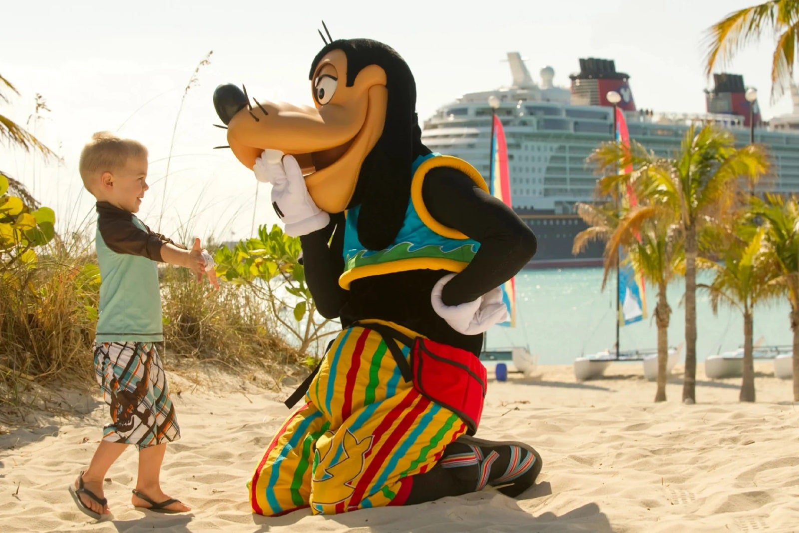 Disney character Goofy kneels in the sand with his hand over his mouth as he interacts with a small child. There's a Disney ship docked in the background.