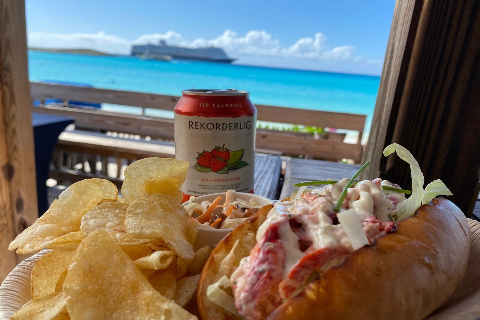 A close-up of a lobster roll with chips and a can of cider with blue sky, water and a ship in the background