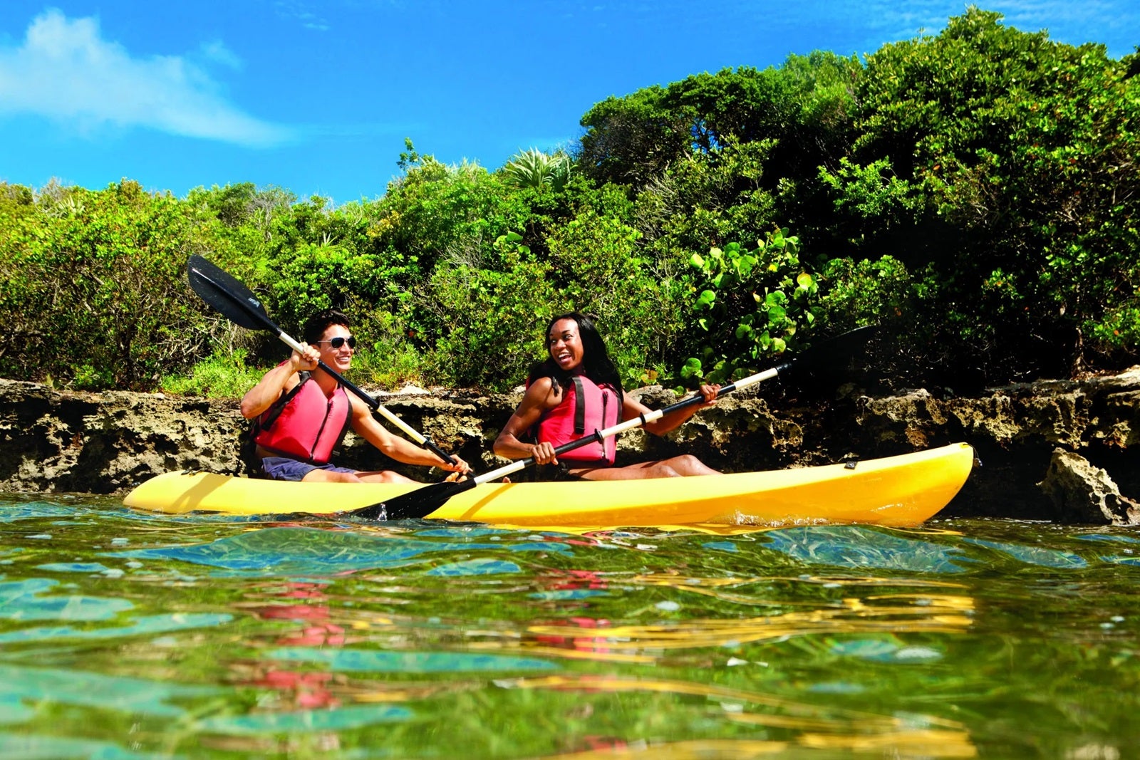 People in red life vests paddle in yellow kayaks with greenery and blue sky in the background.