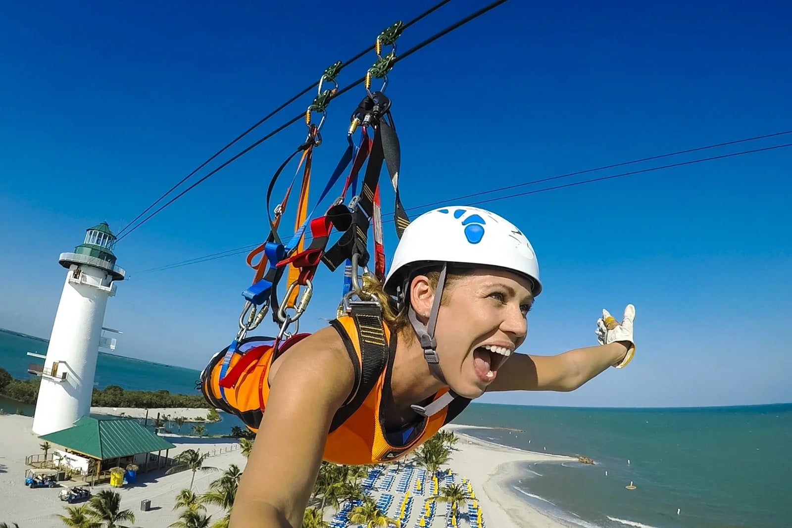 A woman wearing a white helmet takes a selfie as she's supended on a zipline over sand and the ocean with a white lighthouse in the background