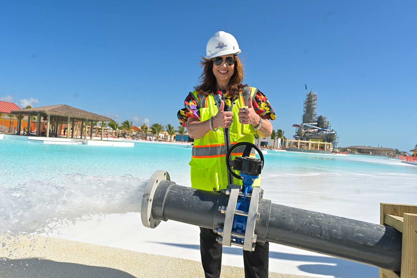 A woman in a yellow contruction vest and white hard hat giving two thumbs up while standing behind a large metal pipe spraying water into a pool