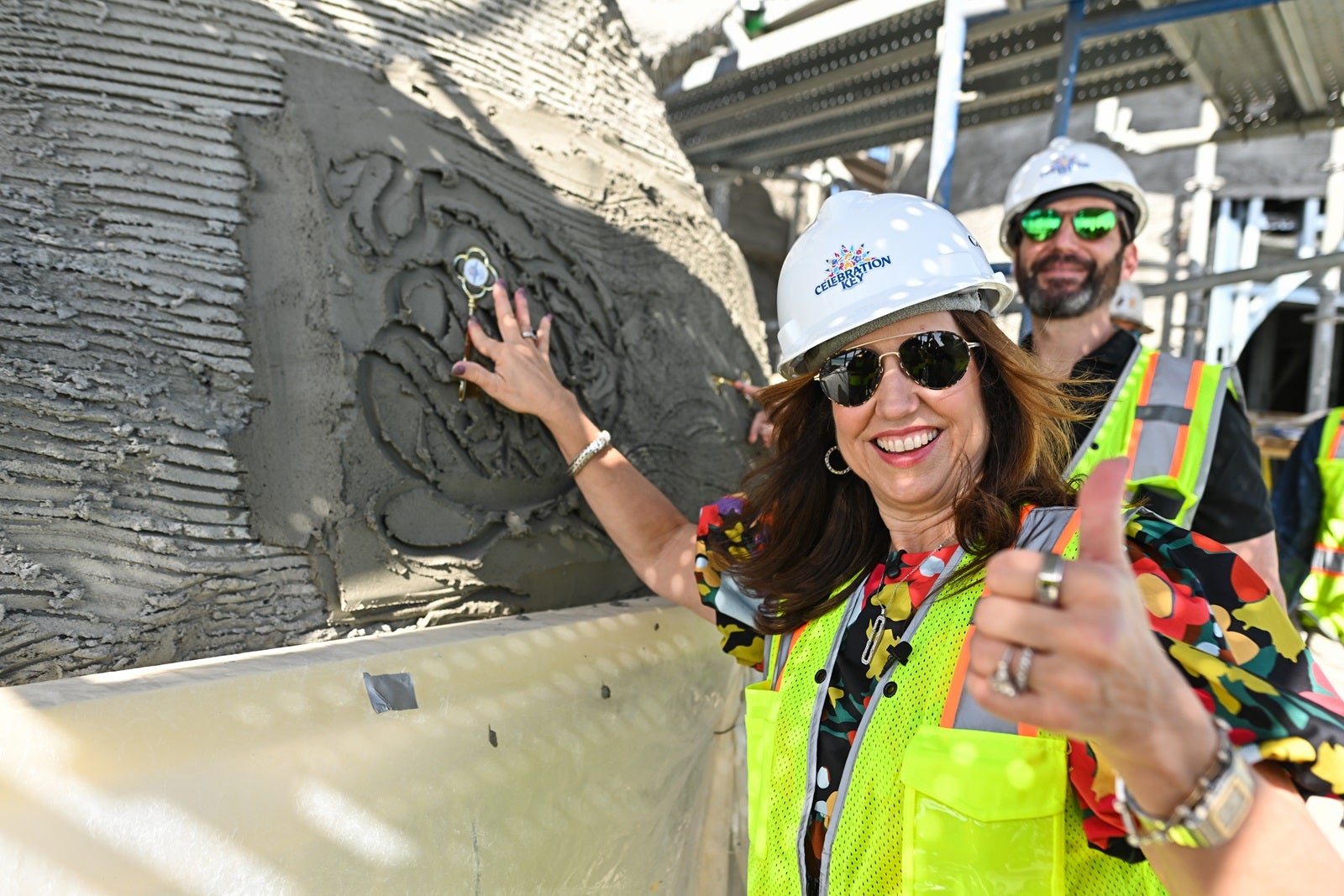 A woman in a yellow construction vest and white hard hat gives a thumbs-up while placing a key into cement as a man in sunglasses smiles behind her