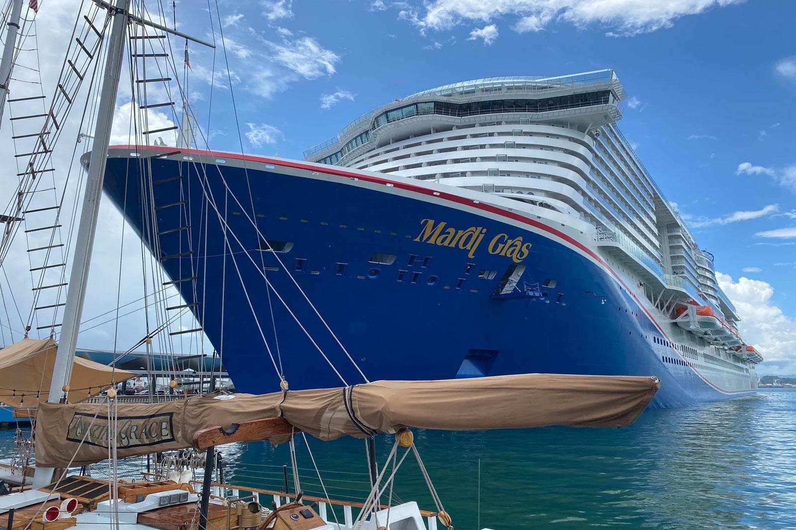 A cruise ship with a blue hull that says "Mardi Gras" docked in front of a masted sailing vessel