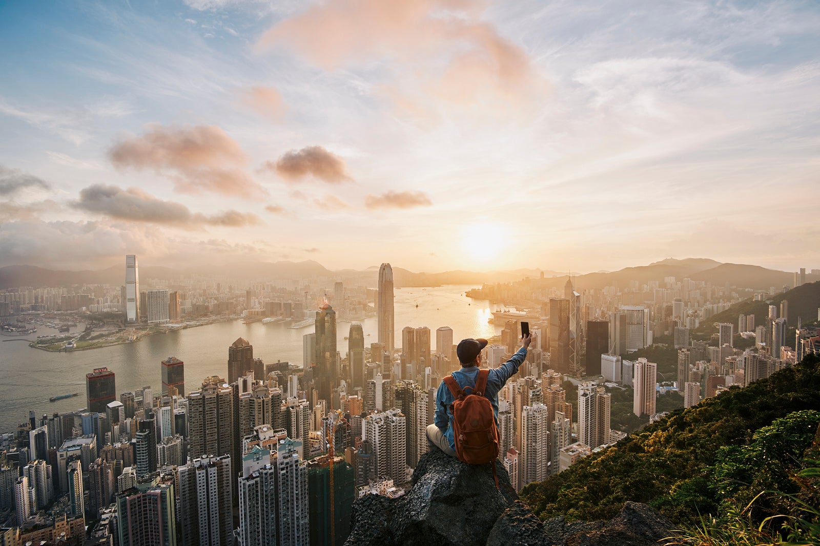 Male tourist sitting on rock in front of Hong Kong skyline at dawn, using mobile phone to take photo