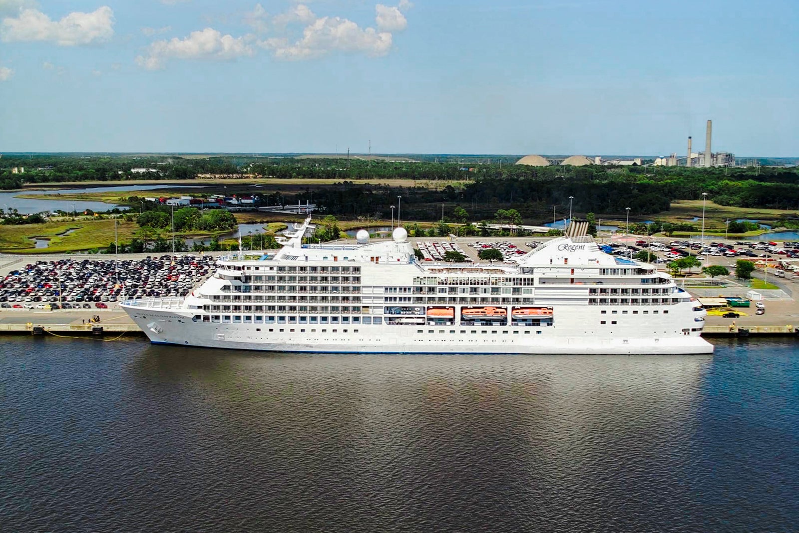 Luxury cruise ship docked with parking lot and factories in the background